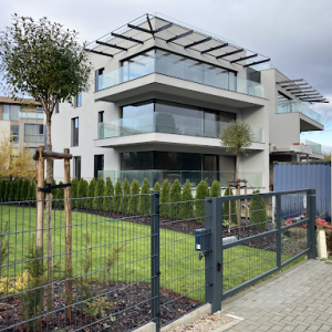 Moderní panelový plot s bránou před novostavbou s prosklenými balkony a upravenou zahradou.